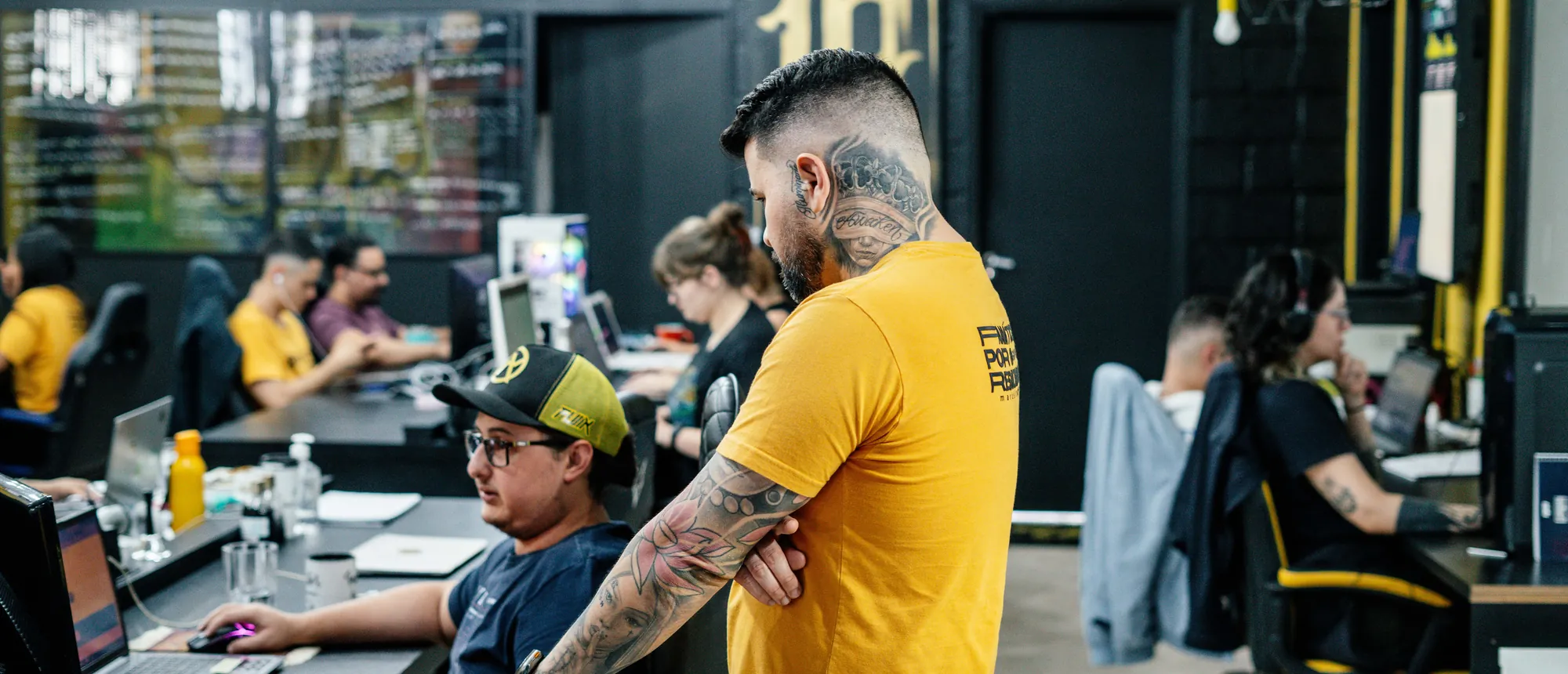 Felipe Jacoto em pé no meio da equipe, acompanhando o trabalho nas mesas do escritório.