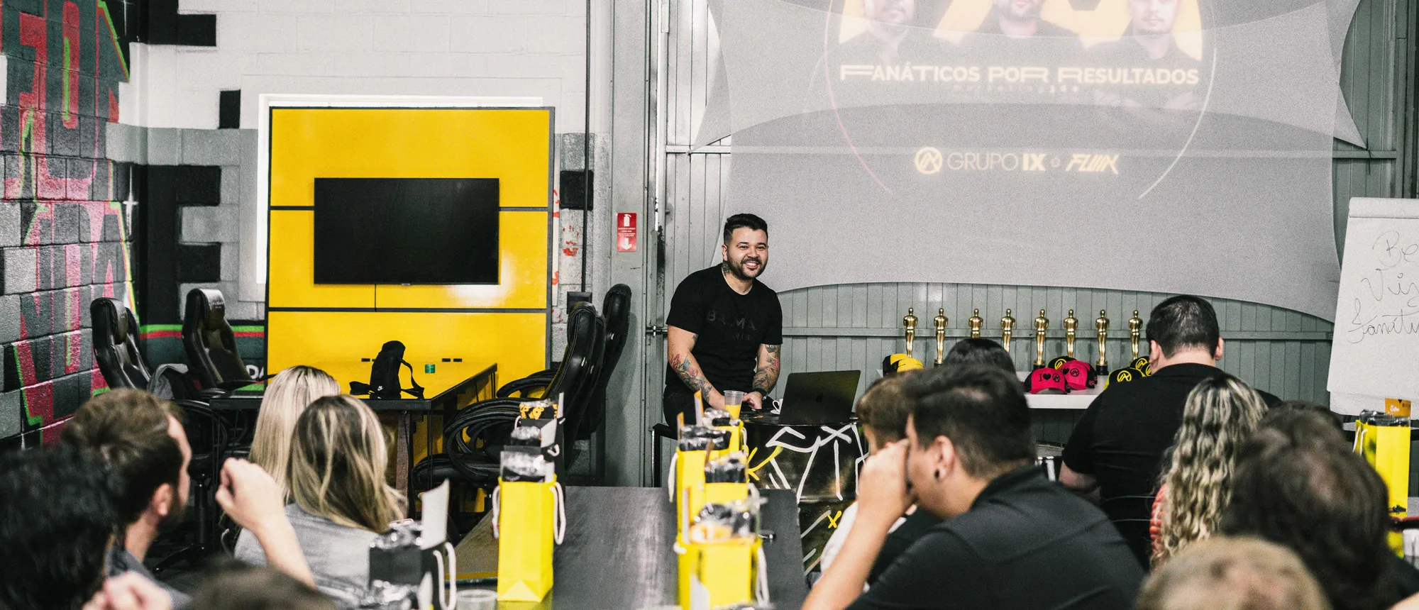 Felipe Jacoto conduzindo uma apresentação para uma sala cheia, com a projeção "Fanáticos por Resultados" ao fundo.